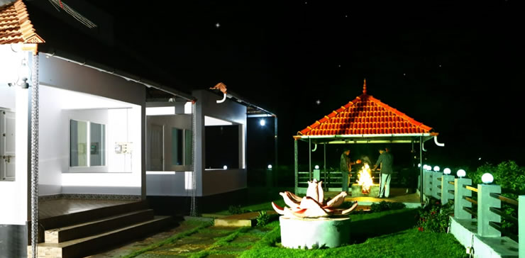 Night view of Ooty estate cottage with guests enjoying a bonfire under a tiled gazebo