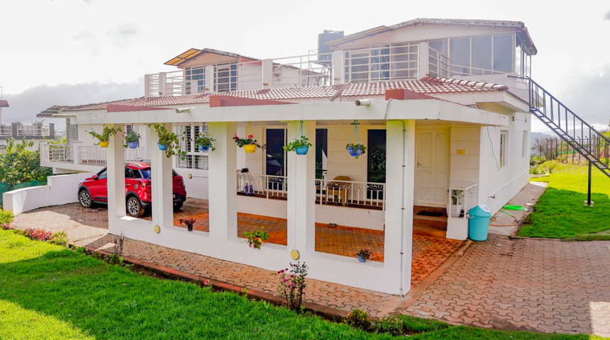 White bungalow with red tiled roof, rooftop terrace, hanging plants, carport with red car, paved walkway, and green lawn