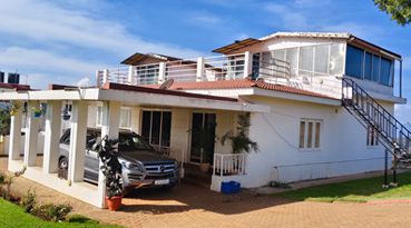 White bungalow exterior in Ooty with covered car parking area, lawn, and rooftop balcony under blue sky